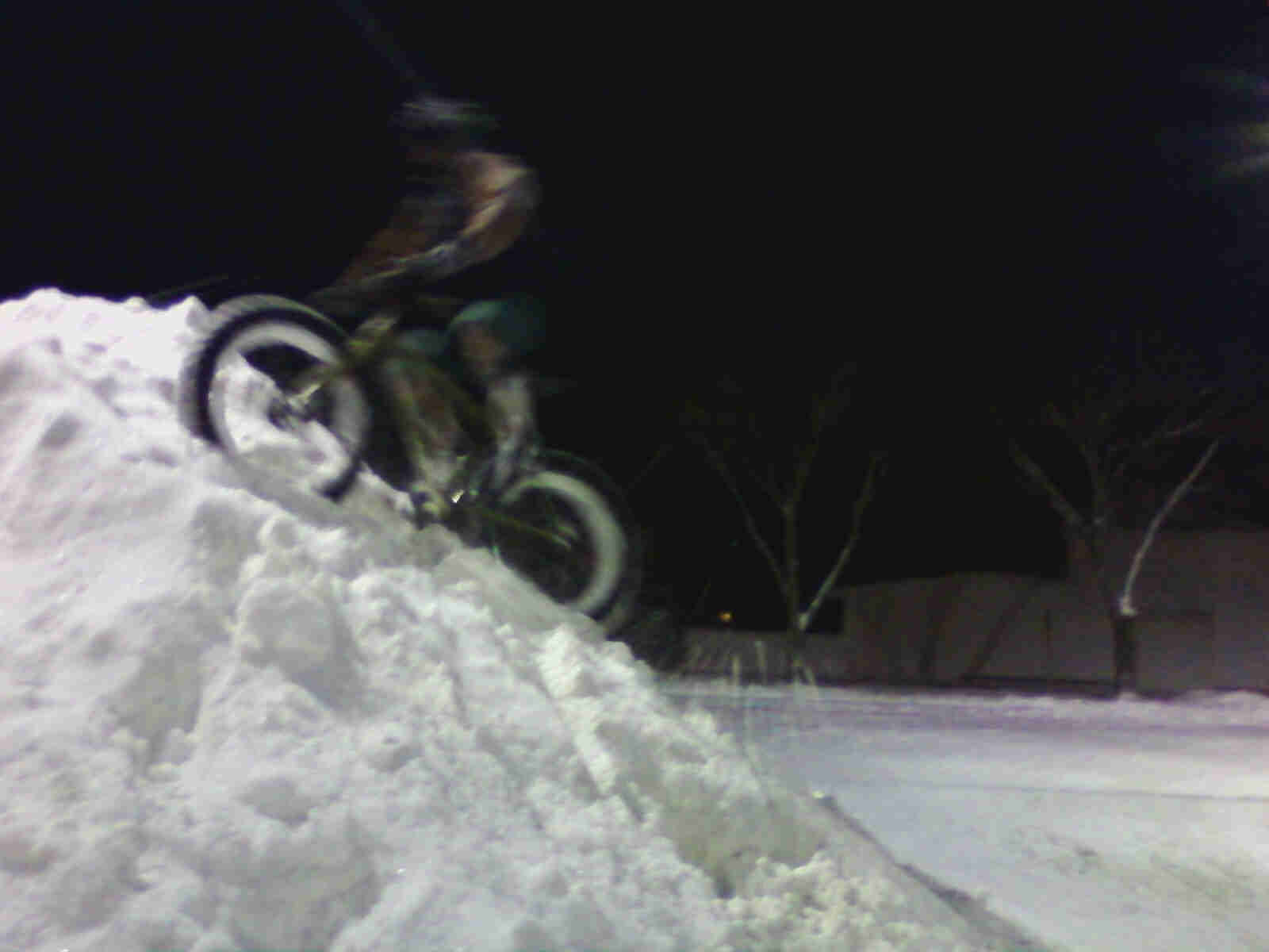 Left side view of a cyclist riding a Surly Pugsley fat bike near the top of a large snow pile, on a parking lot at night